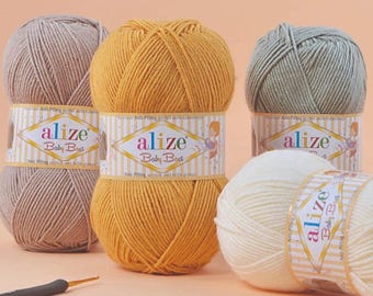 Puede incluir: Primer plano de varios ovillos de lana Alize Baby Best en varios colores: beige, amarillo y verde. Cada ovillo está envuelto con una etiqueta a rayas con el nombre de la marca y una ilustración de dibujos animados. Un ganchillo se encuentra en primer plano.