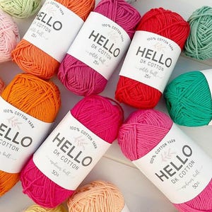 Può includere: Una collezione di gomitoli colorati nei toni dell'arancione, rosa, rosso e verde. Ogni gomitolo è avvolto con un'etichetta bianca che recita "HELLO DK COTTON". Il filato è 100% cotone e disponibile in piccole e medie dimensioni.