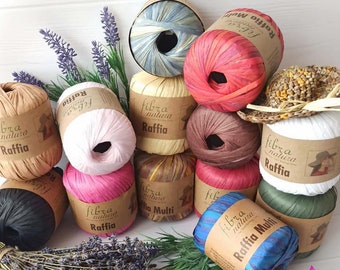 May include: A collection of raffia yarn in various colors, including pink, blue, brown, and white. Each skein is labeled with "fibra natura Raffia" on a brown paper label. A small raffia hat sits atop one of the skeins.