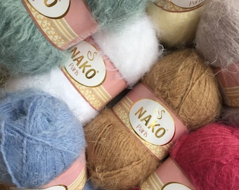 Op de afbeelding: Een verzameling kleurrijke Nako Paris garenbollen. Het garen is verkrijgbaar in verschillende kleuren, waaronder blauw, wit, bruin, roze en groen. Elke bol heeft een roze en gouden label met de merknaam.