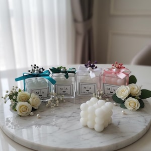 Può includere: Quattro scatole quadrate trasparenti con contenuto bianco, legate con nastri turchese, verdi, bianchi e rosa, sono su un vassoio di marmo. Ogni scatola ha un'etichetta con le iniziali "MA" e una data. Rose bianche e una candela a bolle sono anche sul vassoio.
