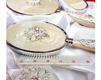 May include: A set of three silver-toned hand mirrors with ornate handles and pink rhinestone accents. The mirrors have a white, textured surface and are displayed on a white satin fabric.
