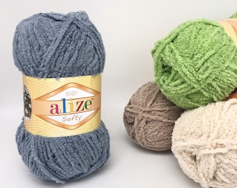 May include: A collection of Alize Softy yarn balls in various colors. The main ball is a soft blue-grey, with the Alize label visible. Other balls are green, brown, and cream-colored, suggesting a range of crafting possibilities.