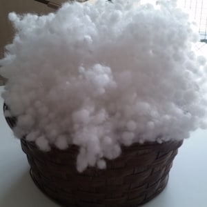 May include: A brown wicker basket filled with white fluffy stuffing. The basket is sitting on a light wood surface.