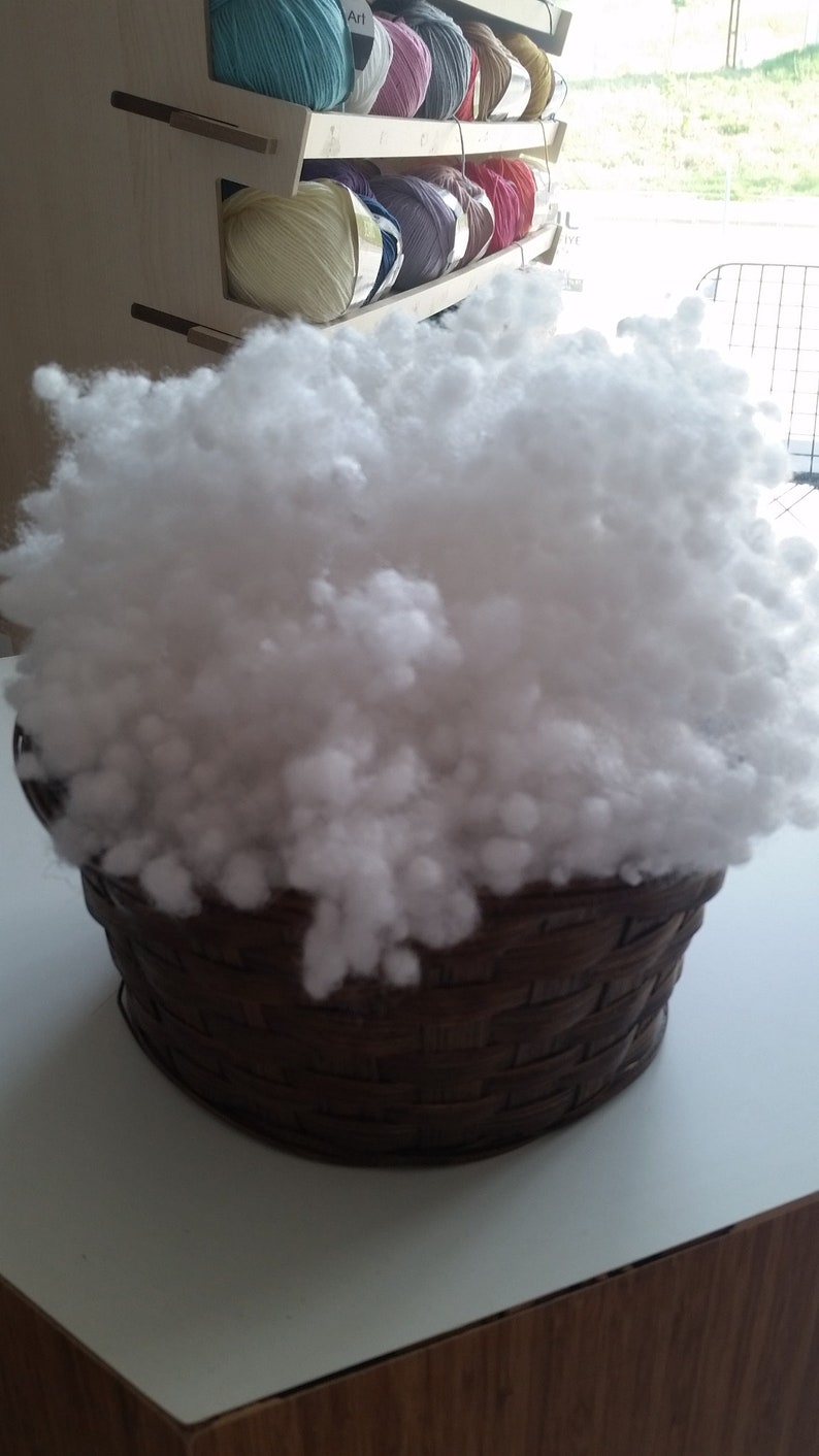 May include: A brown wicker basket filled with white fluffy stuffing. The basket is sitting on a light wood surface.