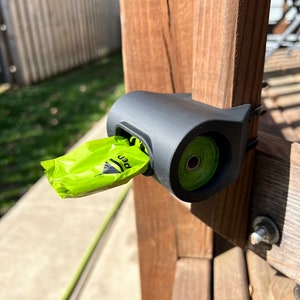 Op de afbeelding: Een zwarte plastic hondenpoepzakdispenser gemonteerd op een houten paal. Een groene rol hondenpoepzakken is zichtbaar in de dispenser. Een groene hondenpoepzak hangt aan de dispenser.