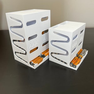 May include: Two white plastic battery organizers with slots for AAA batteries. The larger organizer has a row of batteries in the front slot.