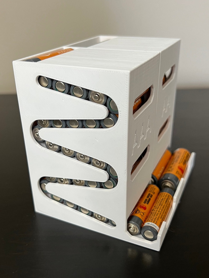 May include: White plastic battery organizer with a zig-zag design to hold AA batteries. The organizer has a separate compartment for AAA batteries.