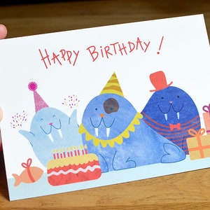 Peut inclure: Carte d'anniversaire avec l'inscription "Happy Birthday!" en rouge. La carte présente des illustrations de trois morses bleus portant des chapeaux de fête et des nœuds papillon, avec un gâteau d'anniversaire et des cadeaux.