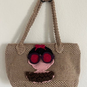 May include: A beige crocheted tote bag with a crocheted face design. The face has pink skin, black hair, red sunglasses, and a brown crocheted mustache.