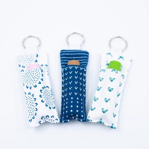 May include: Three fabric keychains with different patterns and contents. One has a pink item, another a brown item, and the third a green item. The fabric patterns include blue dots, blue chevrons, and teal hearts on a white background.