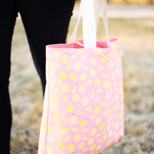 Op de afbeelding: Roze draagtas met een herhaald patroon van gele citroenen en citroenplakjes. De tas heeft witte handvatten en een roze binnenkant. De tas wordt vastgehouden door een persoon die een gele trui en een zwarte broek draagt.