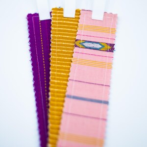 Peut inclure: Trois marque-pages en tissu de couleurs vives : violet, doré et rose, avec des boucles en ruban blanc. Les marque-pages présentent des motifs décoratifs et des bords cousus. Le rose a un motif géométrique.