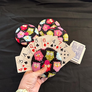 May include: A set of six colourful owl-themed fabric coasters with a hand holding a pack of playing cards. The cards are fanned out and show the numbers 3, 4, 6, and 7.