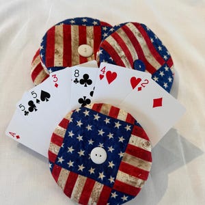 May include: Round fabric card holders with a patriotic American flag design in red, white, and blue. Several playing cards are fanned out, showing the suits and numbers. The holders have a button closure.