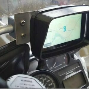 May include: A black motorcycle GPS mount with a sunshade and a built-in screen protector. The mount is attached to a motorcycle handlebar and has a silver metal bracket. The GPS screen shows a map with a blue pin.