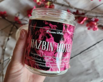 May include: A clear glass jar candle with a label featuring a pink and black smoke pattern. The label text reads Possum Wicks, HAZBIN HOTEL, Bergamot, Sweet Orange, Lavender, Patchouli, Incense, Golden Amber, and Handmade 227g Soy Candle.