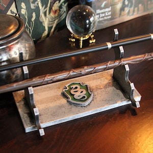 Pode incluir: Um suporte para varinhas de prata com um brasão da casa Slytherin em verde e prata. Duas varinhas são exibidas no suporte.