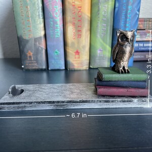 K&ouml;nnte beinhalten: Ein grauer Holzbuchst&uuml;tze mit einem Herz-Ausschnitt. Der Buchst&uuml;tze ist 17 cm lang und h&auml;lt einen Stapel von vier B&uuml;chern. Eine bronzene Eulenfigur sitzt auf den B&uuml;chern. Die B&uuml;cher sind 8,4 cm hoch.
