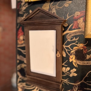 Op de afbeelding: Een donkerbruin houten frame met een witte achtergrond en een sleutelgat. Het frame is gemonteerd op een donkergroen en gouden behang met een patroon van takken en bladeren.