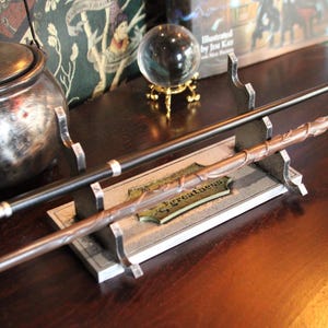 May include: A display stand holding two wands, one black and one brown with a textured vine-like design. The base features a rectangular plaque with the word greatness printed in black lettering. The stand is made of a grey, stone-like material.
