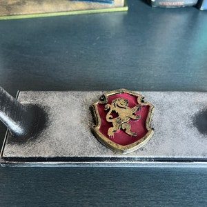 May include: A black wooden stand with a gold lion crest on a red shield. The stand has a distressed finish and is designed to hold a sword or other decorative item.