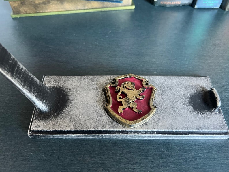 May include: A black wooden stand with a gold lion crest on a red shield. The stand has a distressed finish and is designed to hold a sword or other decorative item.