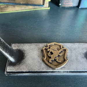 May include: A black wooden stand with a grey distressed finish and a gold lion emblem. The stand has a raised arm for holding a sword or other object.