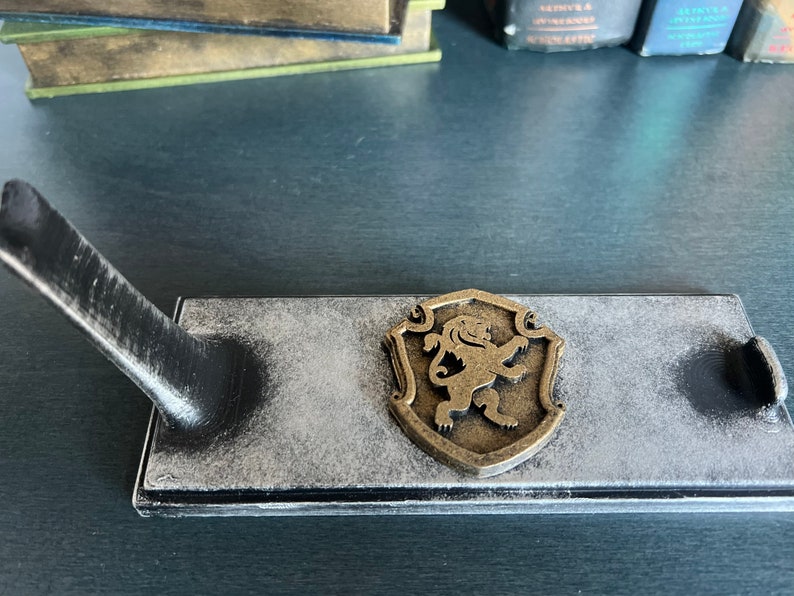 May include: A black wooden stand with a grey distressed finish and a gold lion emblem. The stand has a raised arm for holding a sword or other object.