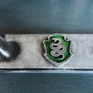May include: A silver-coloured metal stand with a green snake emblem. The stand is designed to hold a wand or other object.
