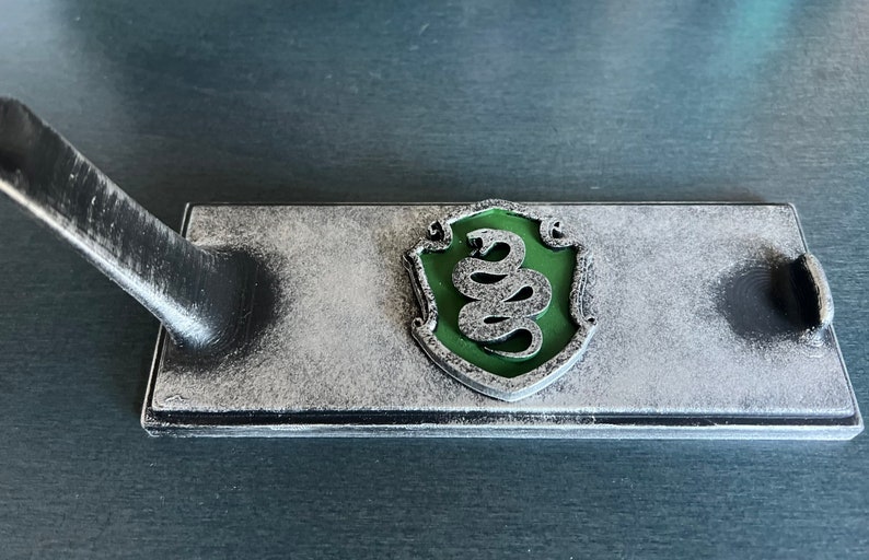 May include: A silver-coloured metal stand with a green snake emblem. The stand is designed to hold a wand or other object.