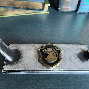 May include: A black wooden stand with a white distressed finish. The stand features a metal badger emblem with a gold finish. The stand is designed to hold a sword or other long object.