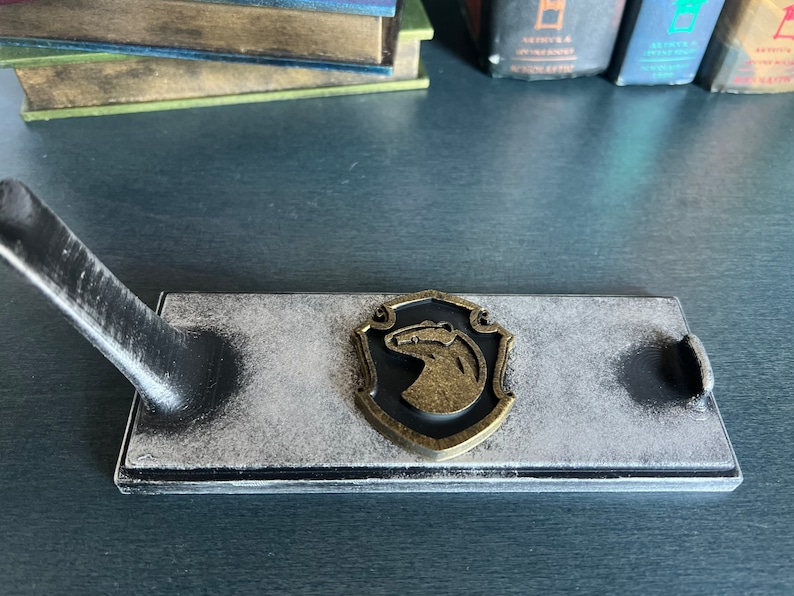 May include: A black wooden stand with a white distressed finish. The stand features a metal badger emblem with a gold finish. The stand is designed to hold a sword or other long object.