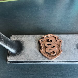 May include: A black and white wooden stand with a copper-coloured snake design in the centre. The stand has a distressed finish and is designed to hold a sword or other decorative object.