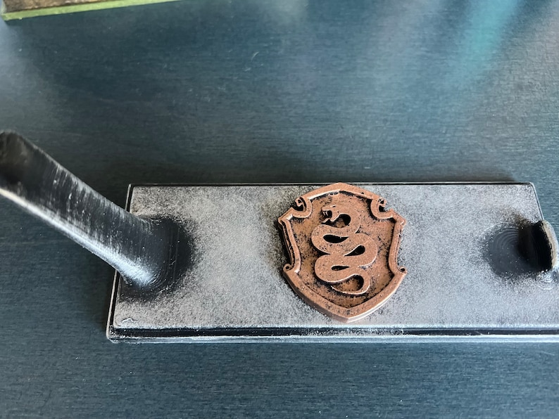 May include: A black and white wooden stand with a copper-coloured snake design in the centre. The stand has a distressed finish and is designed to hold a sword or other decorative object.