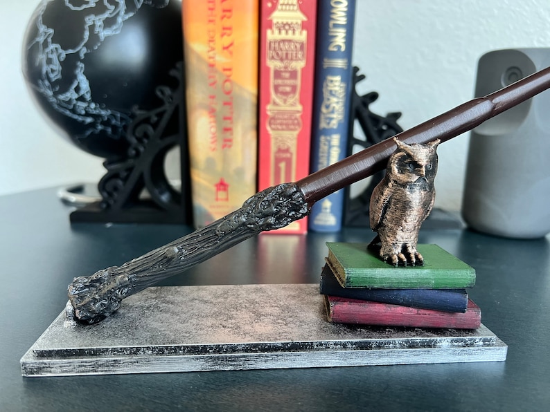 K&ouml;nnte beinhalten: Ein dunkelbrauner Holzstab, der auf einem silbernen Metallsockel mit einem Stapel von drei B&uuml;chern liegt. Eine bronzene Eulenfigur sitzt auf den B&uuml;chern.