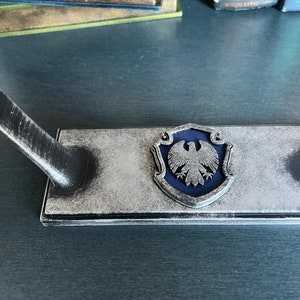 May include: A silver and black decorative stand with a blue eagle crest. The stand has a raised edge and a small, curved arm.