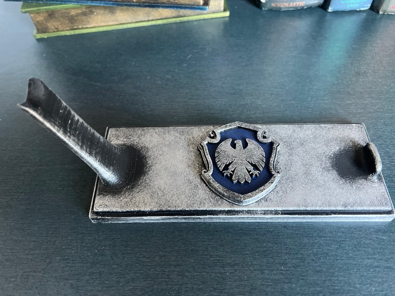 May include: A silver and black decorative stand with a blue eagle crest. The stand has a raised edge and a small, curved arm.