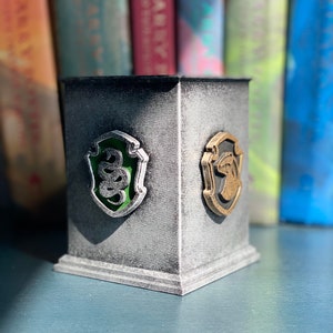 May include: A silver and grey pencil holder with a green and silver Slytherin house crest and a gold and black Hufflepuff house crest.