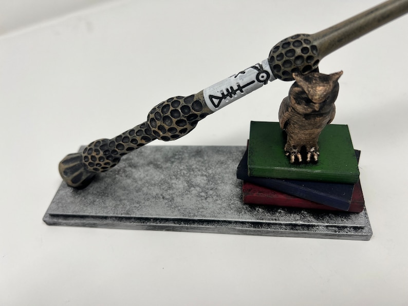 K&ouml;nnte beinhalten: Ein brauner und goldener Zauberstab mit einem wei&szlig;en Fischsymbol darauf liegt auf einem silbernen St&auml;nder. Eine kleine goldene Eulenfigur sitzt auf einem Stapel gr&uuml;ner, blauer und roter B&uuml;cher.