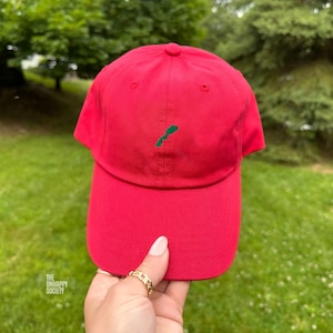 Könnte beinhalten: Eine leuchtend rote Baseballkappe mit einer kleinen, grün gestickten Landkarte von Marokko in der Mitte der Vorderseite. Die Kappe hat eine strukturierte Krone, einen gebogenen Schirm und einen Knopf oben. Der Text THE UNHAPPY SOCIETY ist zu sehen.