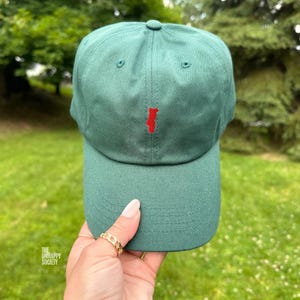 Könnte beinhalten: Eine waldgrüne Baseballkappe mit einer kleinen, aufgestickten roten Silhouette von Portugal in der Mitte des Vorderteils. Die Kappe hat eine strukturierte Krone, einen gebogenen Schirm mit sichtbaren Nähten und einen Knopf oben in der Mitte.