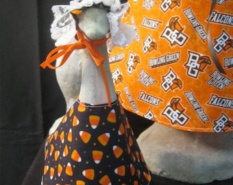 May include: A bowling pin dressed in a black candy corn print apron and a white lace bonnet with an orange ribbon. Another pin is partially visible, wearing an orange fabric with the text "BOWLING GREEN FALCONS".