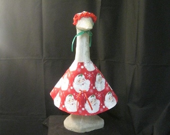 May include: A white goose wearing a red Santa Claus themed dress with a red and white Santa Claus print and a red ruffled hat with a green ribbon.