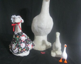 May include: A collection of four goose figurines of varying sizes. One large white goose wears a black dress with a Santa Claus print, a red ribbon, and a white lace bonnet. Beside it are two plain white goose statues and one small white goose with orange feet.