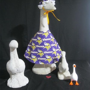 May include: A large white goose statue wearing a purple dress with a yellow and white Vikings logo pattern and a matching bonnet with a yellow ribbon. Three additional goose statues of varying sizes are present, including one small white goose with orange feet.
