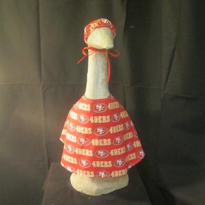 May include: A white ceramic goose figurine wearing a red and gold 49ers themed hat and cape. The cape is covered in the words "49ers" repeated multiple times.