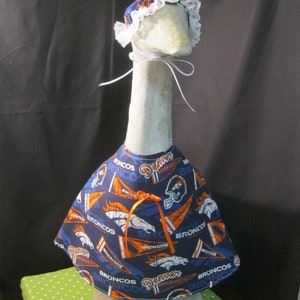 May include: A white goose figurine wearing a blue and orange Denver Broncos themed fabric dress with a white lace bonnet. The dress has the words "Denver Broncos" printed on it.