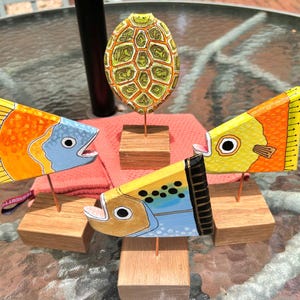 May include: A collection of four hand-painted wooden sculptures featuring three fish and one turtle shell, each mounted on a square wooden base with a thin metal rod. The fish are painted in bright yellow, orange, and blue, while the turtle shell is green.