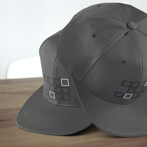 May include: Two gray baseball caps with a black and white geometric design on the front. The caps are on a wooden surface.